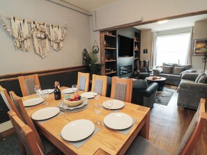 Uma sala de jantar com mesa de madeira posta para seis pessoas, rodeada por cadeiras. Há uma televisão ao fundo e uma área de estar com sofás.
