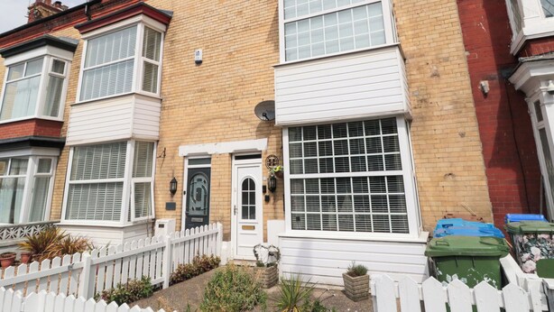 A two-story residential building with a white front door, a small garden with potted plants, and a white picket fence. - Memory Maker (Bridlington)