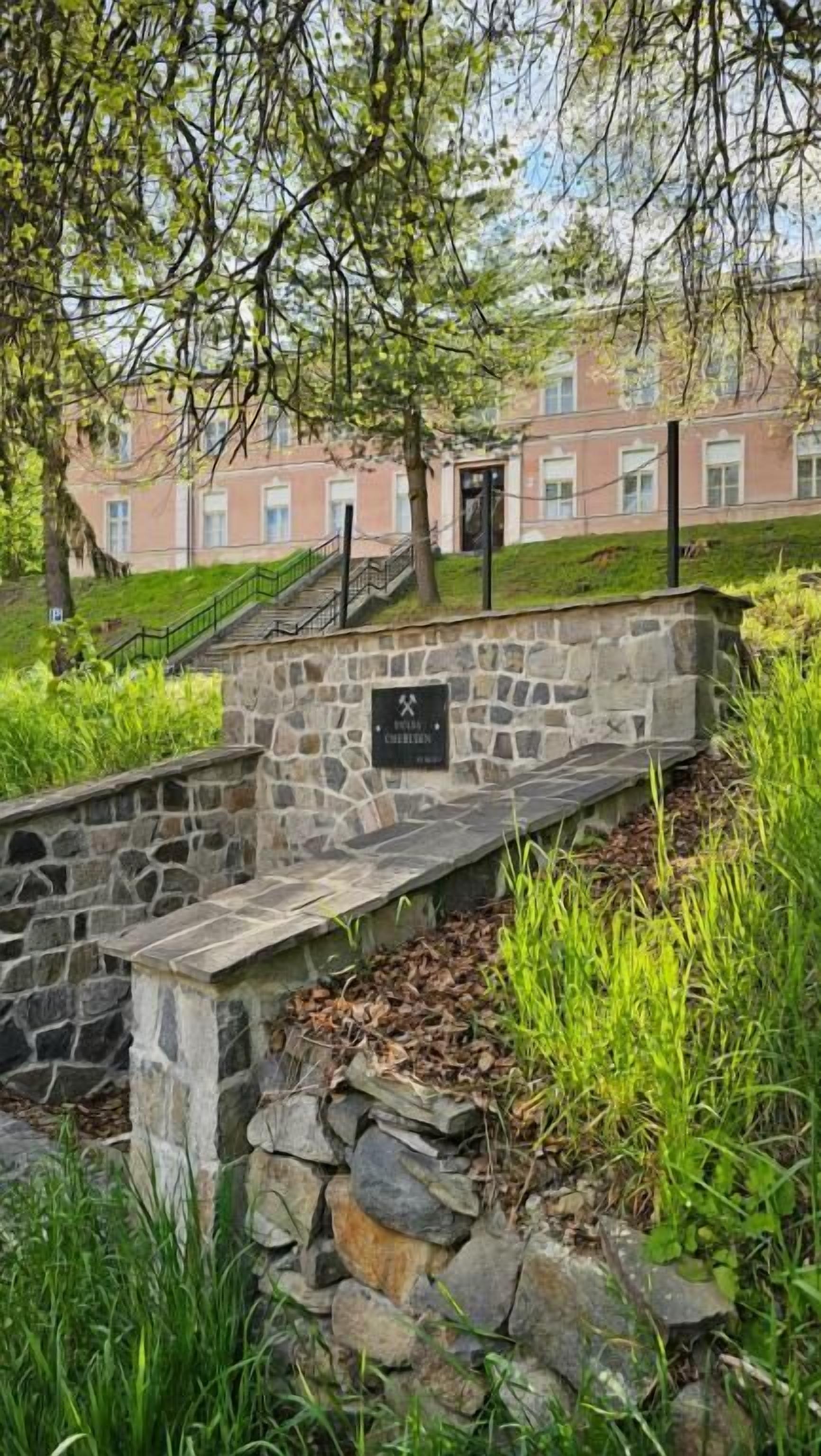 A stone bench with a plaque, surrounded by greenery and a building in the background.