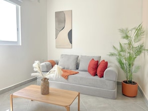 Una sala de estar con un sofá gris, una mesa de centro de madera, un jarrón con plumas, una planta en maceta y un cuadro abstracto grande en la pared.