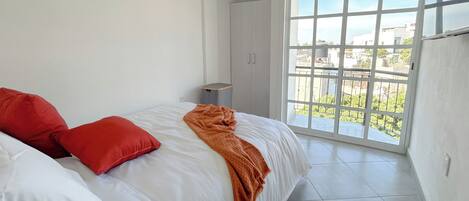 A bedroom with a bed, red pillows, a white blanket, a window with a view, and a small table.