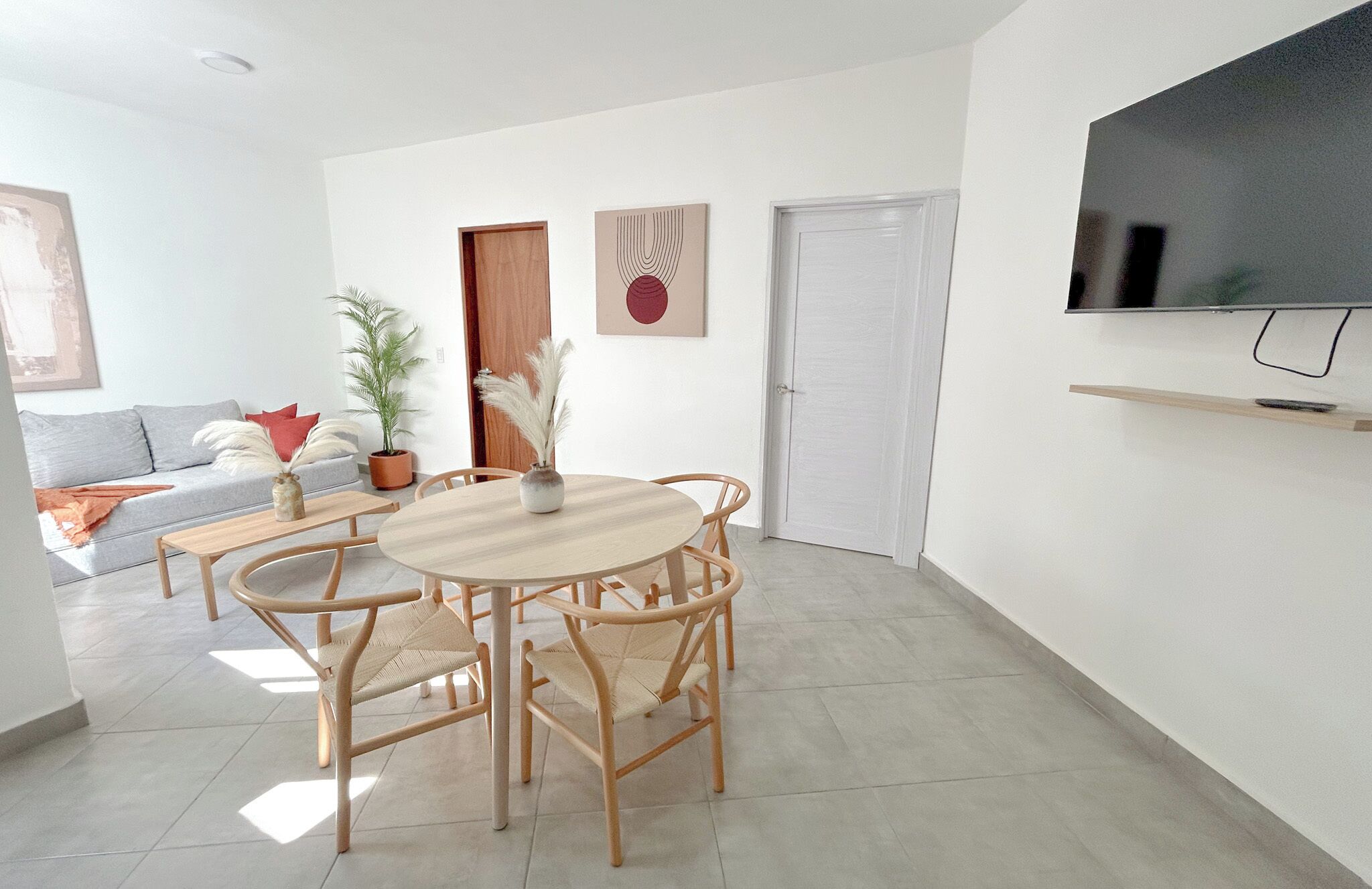 Ein modernes Wohnzimmer mit einem runden Holztisch, vier Stühlen, einer Couch und einem an der Wand befestigten Fernseher, sowie einer Tür.