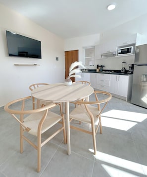 A modern kitchen with white cabinetry, a flat-screen TV mounted above the counter, and a dining area with a round table and wicker chairs. - El Pitillal (Puerto Vallarta)