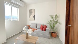 Een woonkamer met een grijze bank, een houten salontafel, een kamerplant en een raam met uitzicht.