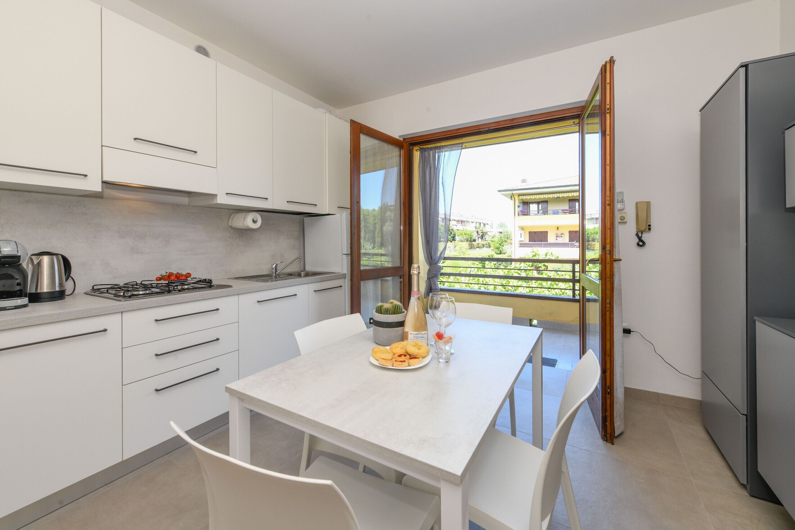 A modern kitchen with white cabinets, a dining table with chairs, a small balcony with a view of buildings, and a refrigerator.