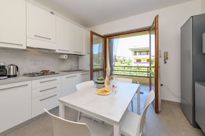 Eine moderne Küche mit weißen Schränken, einem Esstisch mit Stühlen, einem kleinen Balkon mit Blick auf Gebäude und einem Kühlschrank.