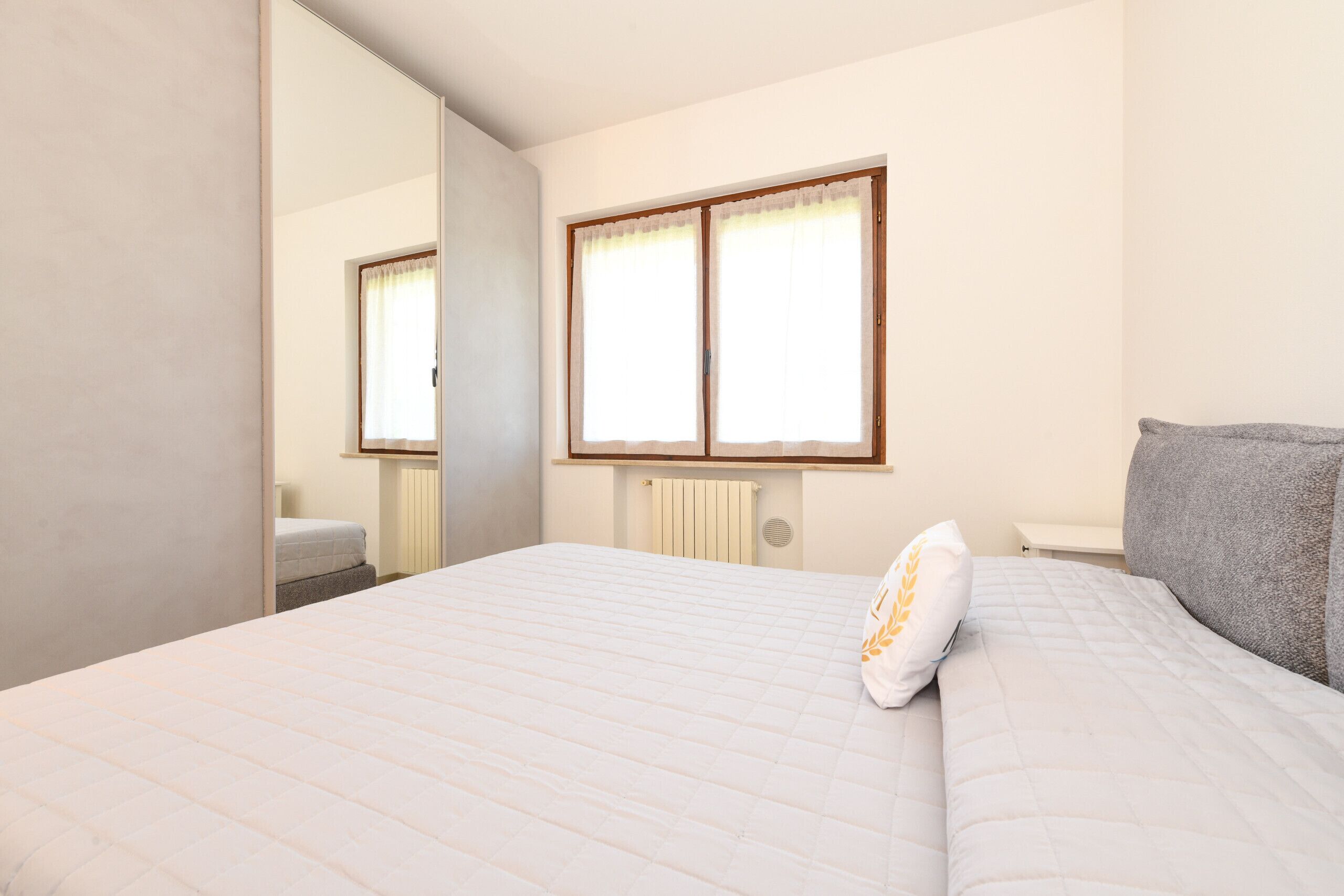 A bedroom with a bed, a window with curtains, a radiator, and a mirror.