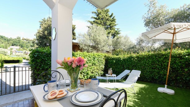 An outdoor patio with a table set for two, a flower vase, and a white umbrella. - MGH- AMAI Small Holiday Cottage (Manerba del Garda)