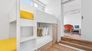 Une pièce moderne et bien éclairée, avec un lit superposé, une porte en bois et une vue sur un espace de vie comprenant un canapé et un fauteuil.