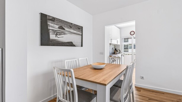 A dining area with a wooden table, white chairs, and a bowl. There is a kitchen visible in the background. - Acolhedor T2 em Armação de Pêra by Lovelystay (Armacao de Pera)