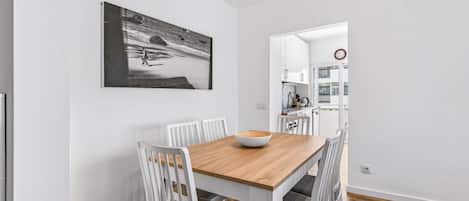 A dining area with a wooden table, white chairs, and a bowl. There is a kitchen visible in the background.