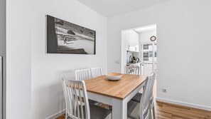 Un coin repas avec une table en bois, des chaises blanches et un saladier. On aperçoit une cuisine à l’arrière-plan.