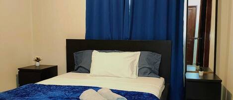 A bedroom with a bed, a nightstand, an air conditioning unit, a mirror, and blue curtains.
