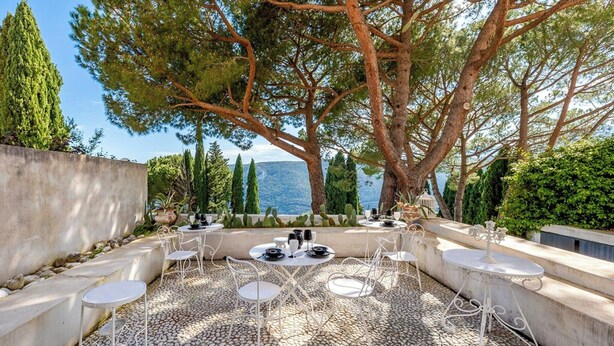 Un área de comedor al aire libre con mesas y sillas redondas, rodeada de árboles y vista al mar.