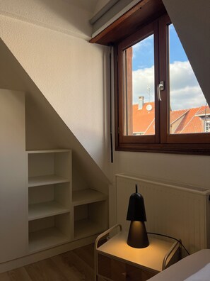 A room with a sloped ceiling, a window with a view of buildings, a radiator, and a chair with a lamp.
