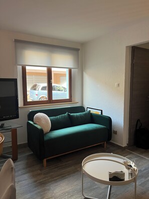 A living room with a green sofa, a small round table, a TV, and a window with a view of parked cars.