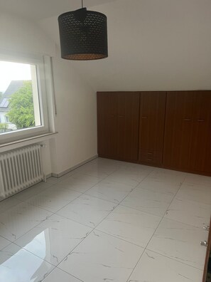 Una habitació amb un radiador, una finestra, un armari de fusta i una làmpada penjant.