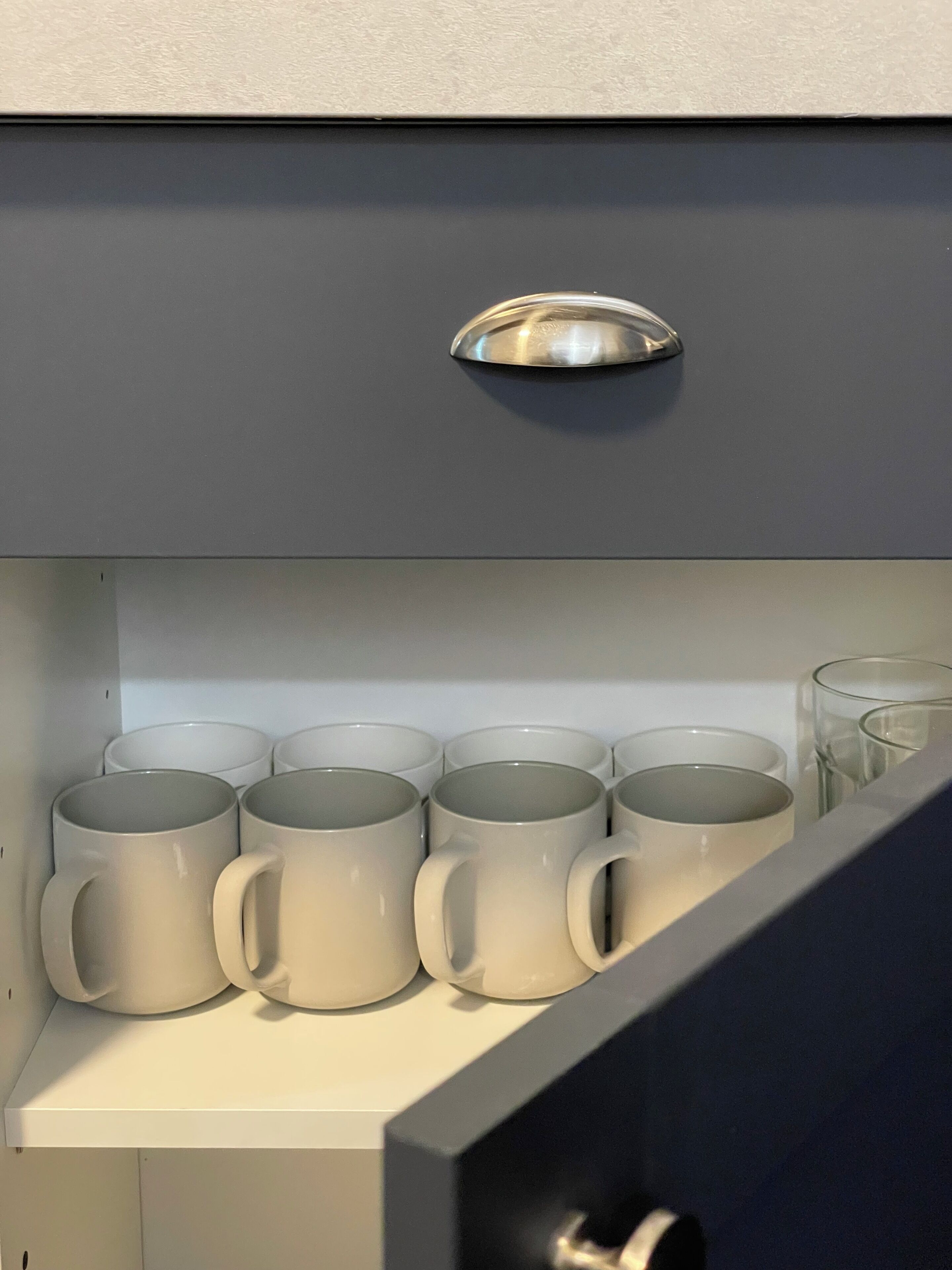 Ein Küchenschrank mit einer Metallstange und einer Ablage mit sechs weißen Tassen und zwei klaren Gläsern.