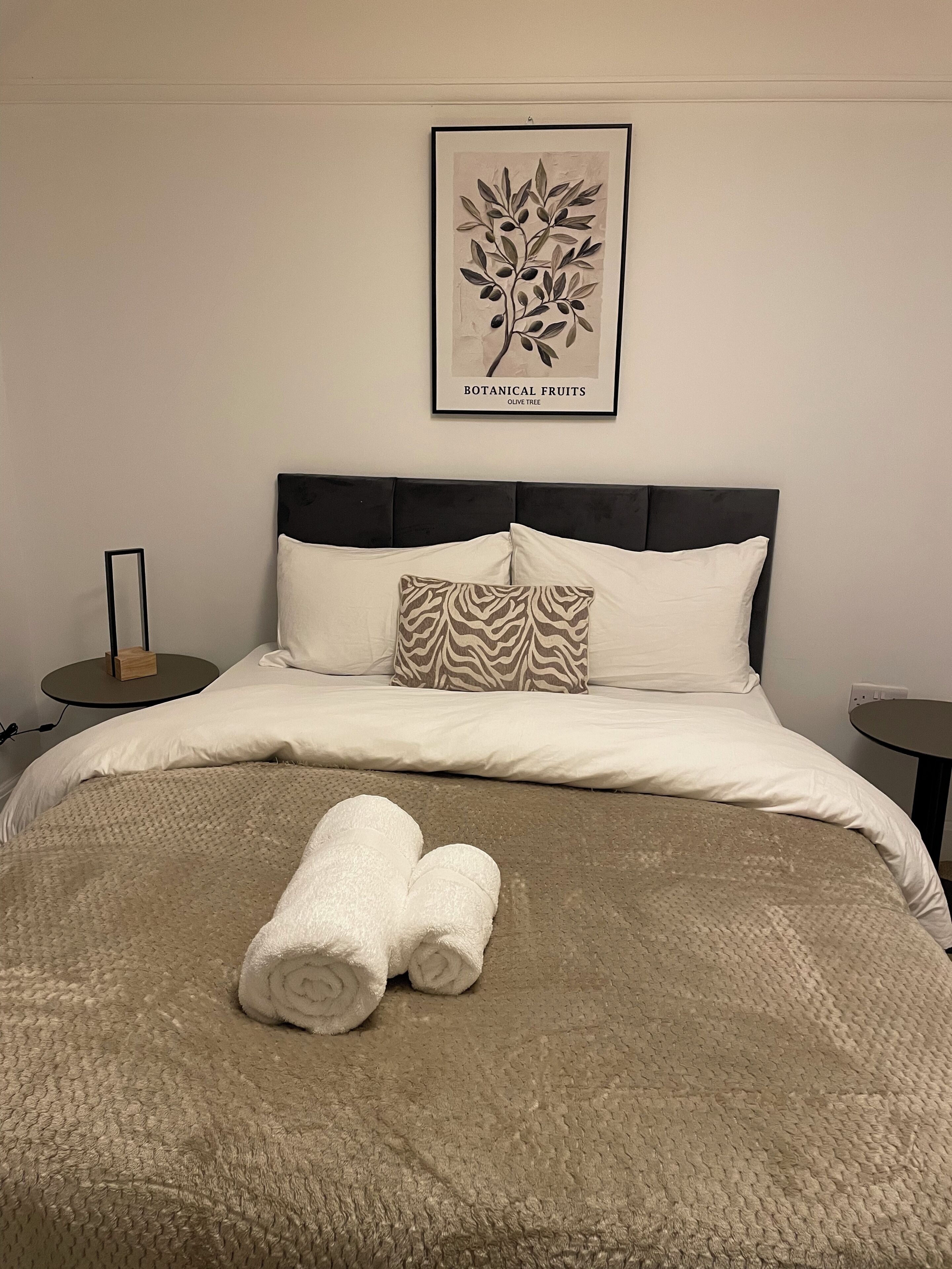 A neatly made bed with a beige blanket, two rolled towels, a zebra-patterned pillow, and a framed botanical print on the wall.