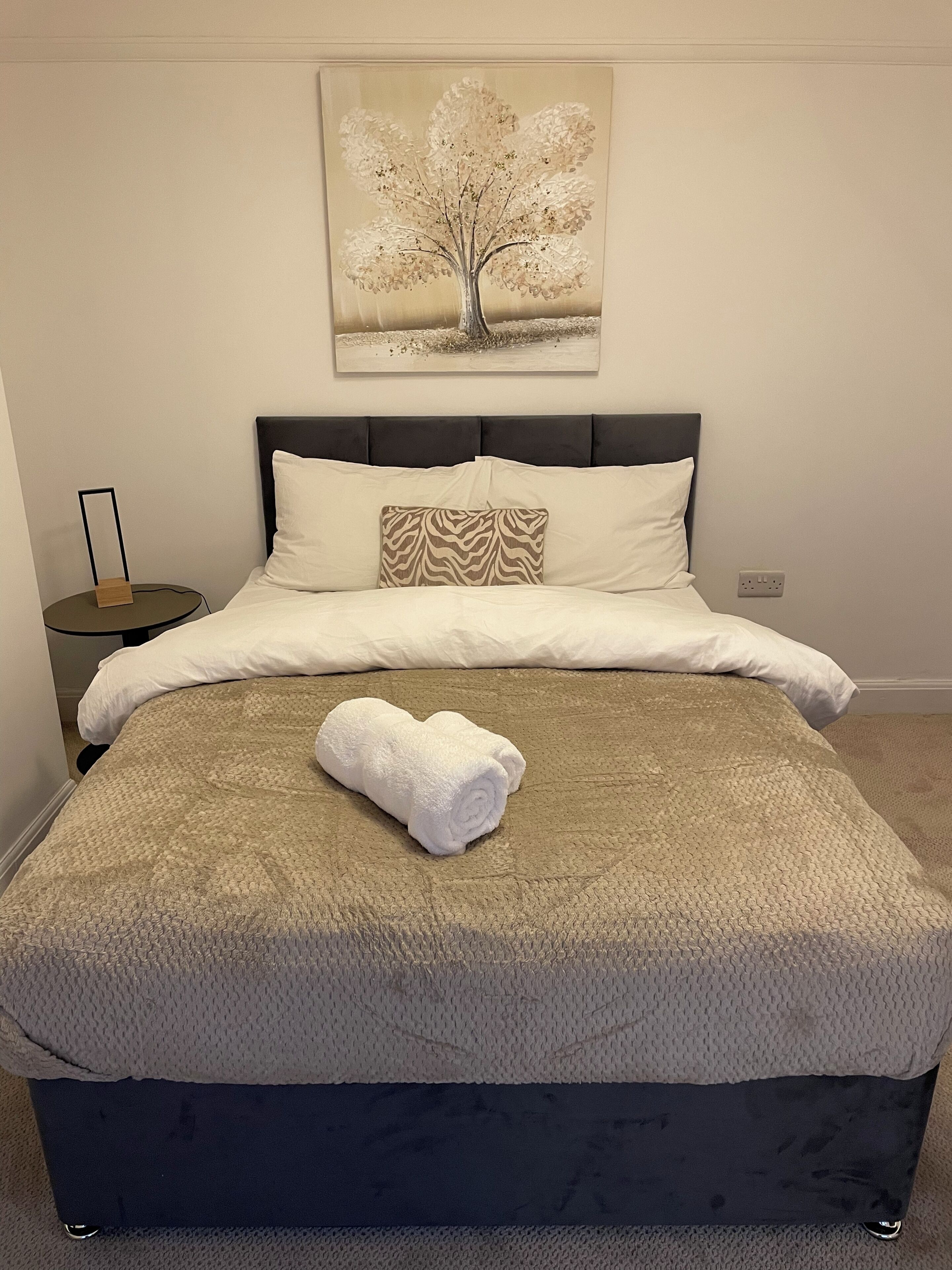 A neatly made bed with a rolled white towel, a zebra-patterned pillow, and a tree-themed artwork on the wall.