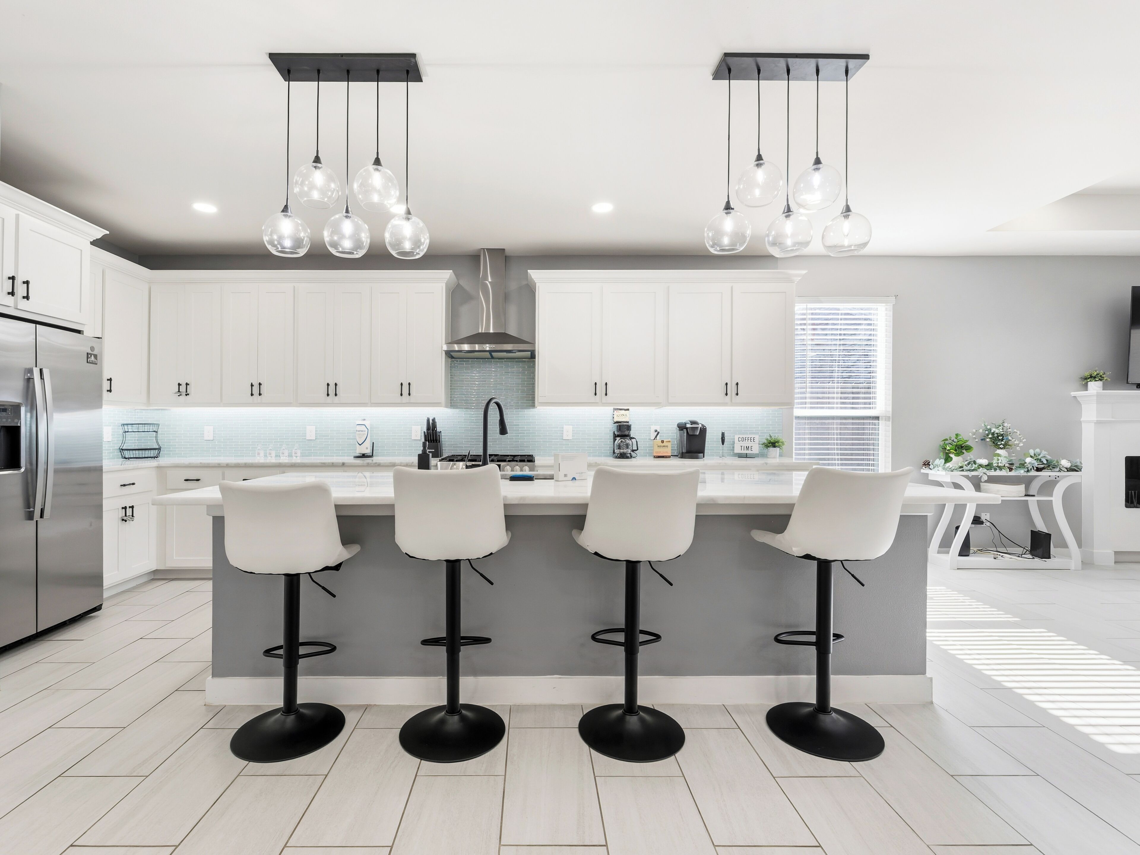 Une cuisine moderne avec des meubles blancs, un réfrigérateur en acier inoxydable, une cuisinière à gaz et trois tabourets de bar.