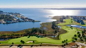 Luftaufnahme eines Küsten-Golfplatzes mit Wasser, Häusern und blauem Himmel.