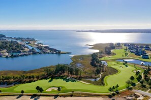 Luftaufnahme eines Küsten-Golfplatzes mit Wasser, Häusern und blauem Himmel.