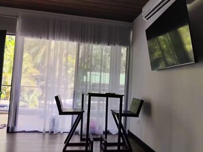 A room with a large window, a TV mounted on the wall, and a table with chairs. - Duke's Place (Quepos)