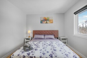 A bedroom with a bed, a nightstand, a lamp, a painting, and a window.