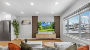 Ein modernes Wohnzimmer mit Fernseher, großem Fenster und Küchenbereich inklusive Kühlschrank.