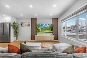 A modern living room with a flat-screen TV, a large window, and a kitchen area with a refrigerator.