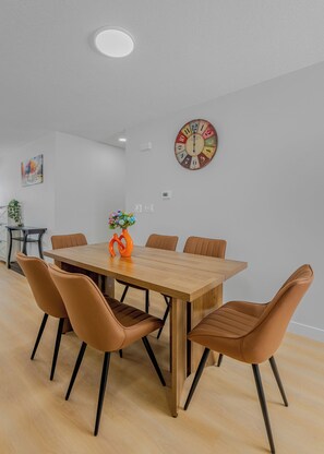 A dining area with a wooden table, six brown chairs, a colorful clock, and a vase with flowers.