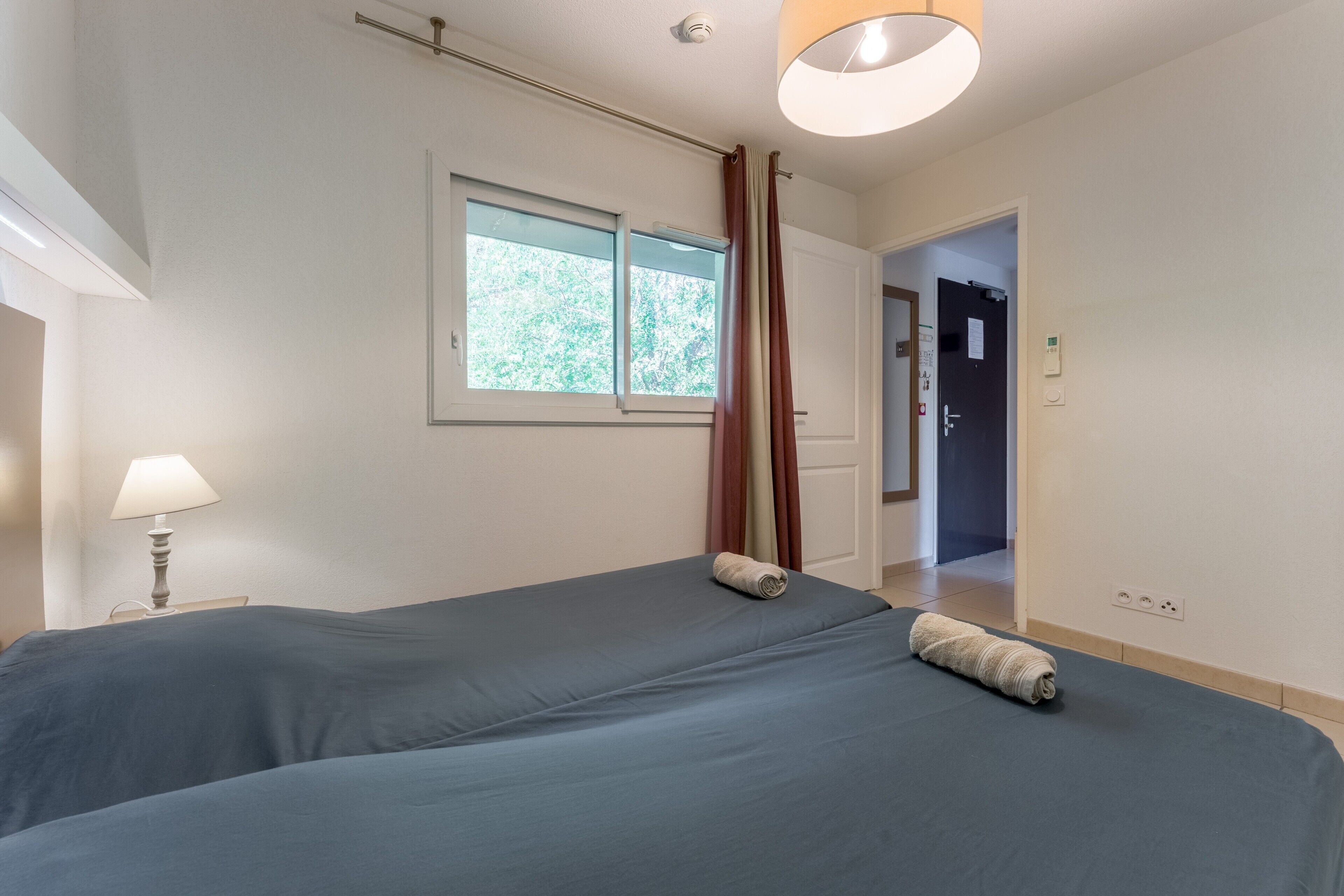 A bedroom with two beds, a window, a lamp, and a door leading to another room.