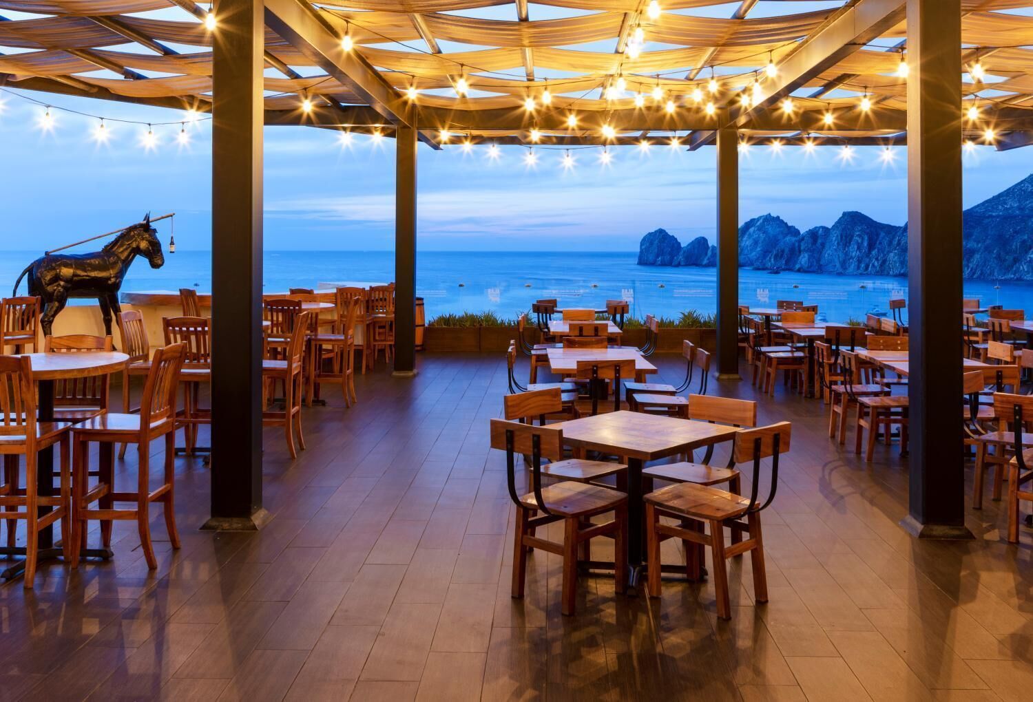 An outdoor dining area with wooden tables and chairs, string lights, and a view of the sea and mountains.