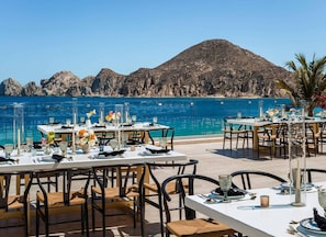 Un área de comedor al aire libre con mesas preparadas para dos, con vistas a un mar azul cristalino y un paisaje montañoso.