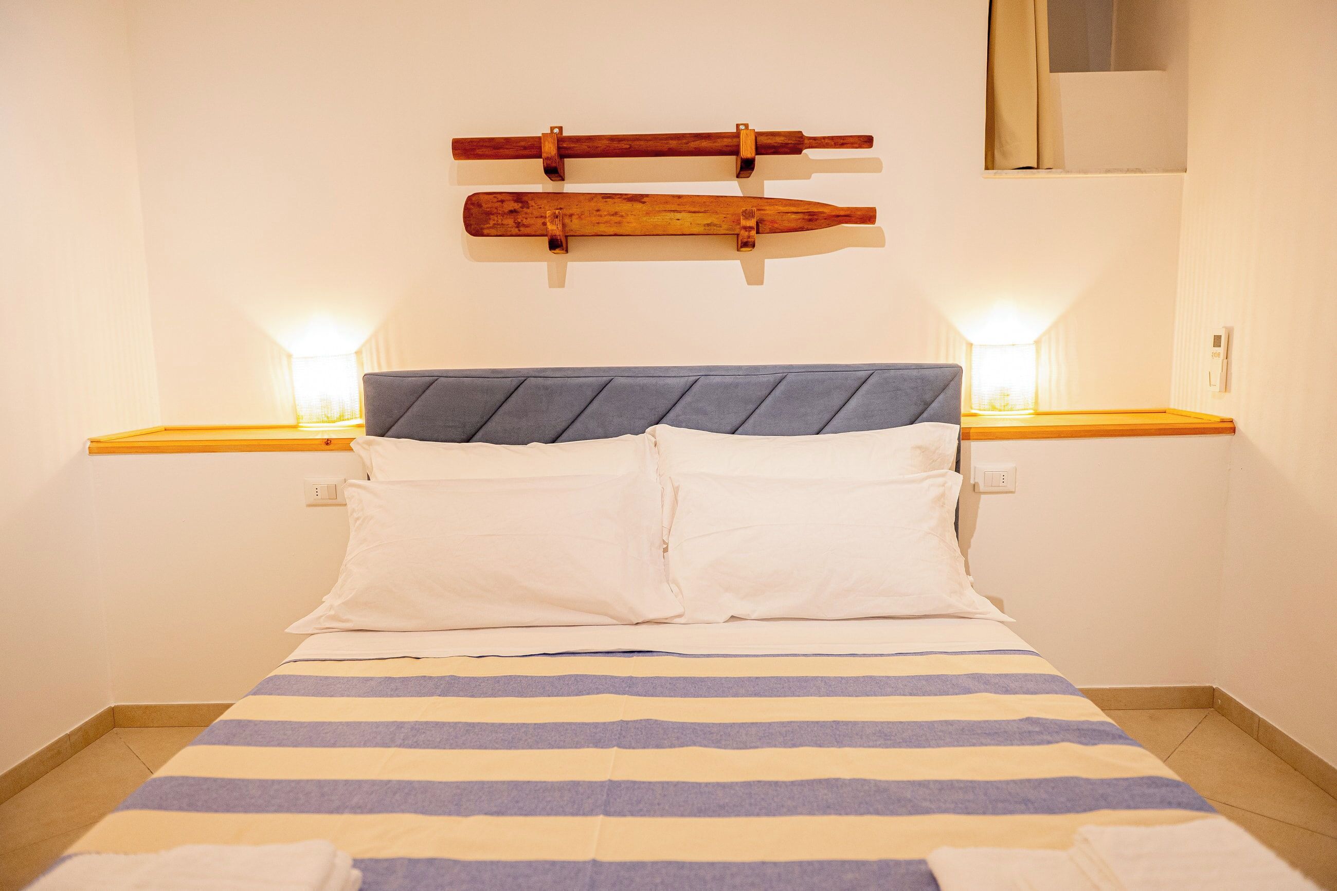 Een bed met een gestreept dekbedovertrek, twee nachtkastjes met lampen, en een houten object aan de muur boven het bed.