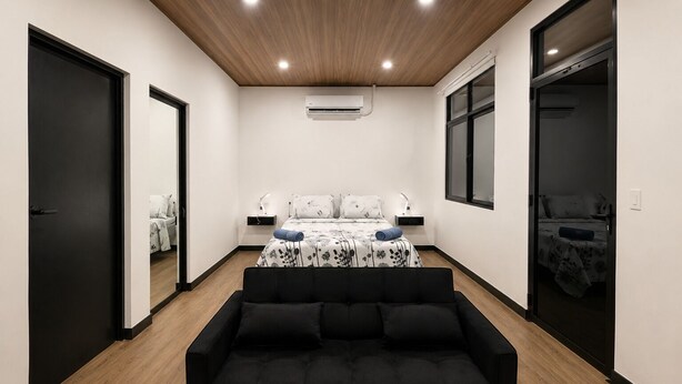 Une chambre moderne avec un canapé noir, un lit recouvert d’une literie à motifs floraux, une table de chevet avec une lampe et une fenêtre.