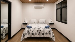 Une chambre avec un plafond en bois, un appareil de climatisation, un lit avec une literie à motifs floraux, deux tables de chevet avec des lampes et une fenêtre avec des stores.