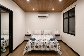 A bedroom with a wooden ceiling, an air conditioning unit, a bed with floral bedding, two bedside tables with lamps, and a window with blinds.
