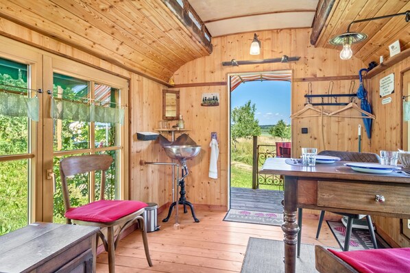 A wooden interior with a dining table, chairs, a hanging light fixture, and a view through the door. - Circus Wagon in Ostrach – Unique Caravan in the Orchard (Ostrach)