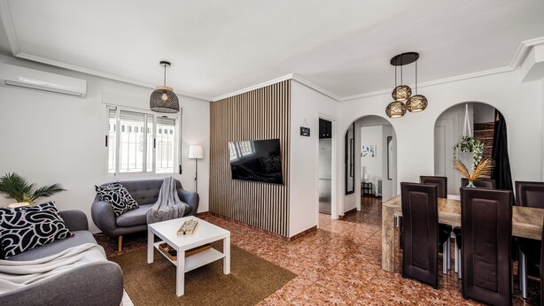 Ein modernes Wohnzimmer mit Flachbildfernseher, grauem Sofa, weißem Couchtisch und einem Essbereich mit dunklen Stühlen und einem Holztisch.