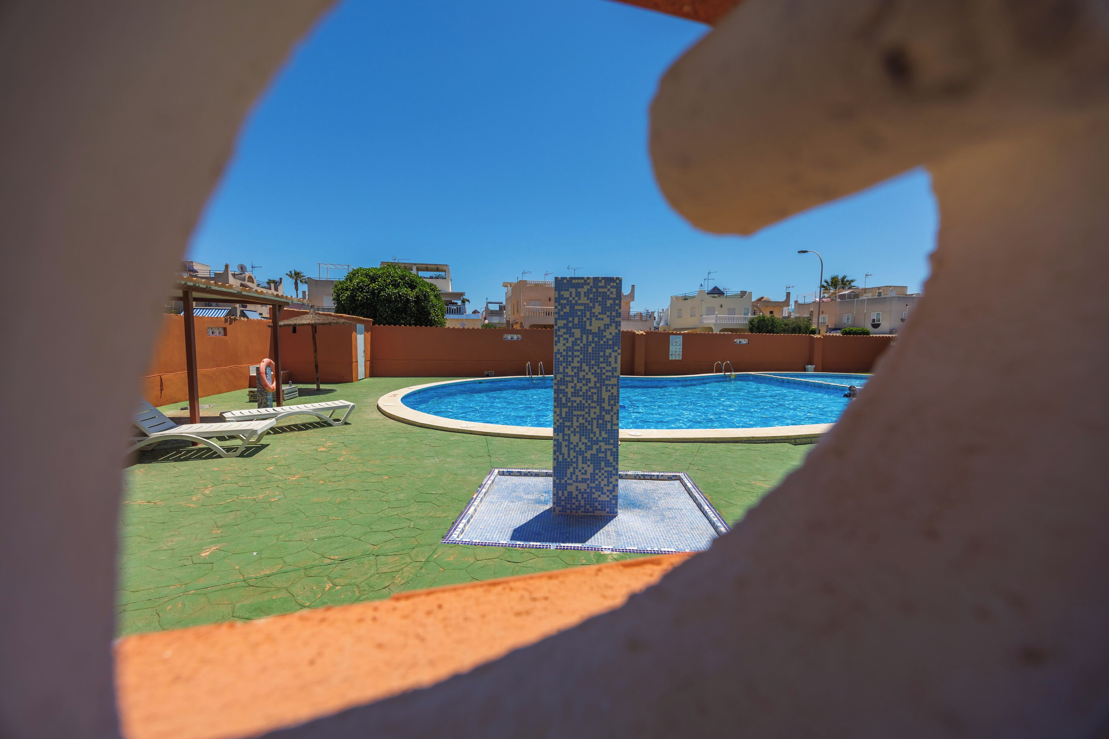 Une piscine agrémentée d’une colonne recouverte de carreaux, d’une chaise longue et offrant une vue sur les bâtiments en arrière-plan.