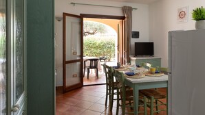 A dining area with a table set for two, a television on a stand, and a door leading to an outdoor area. - Casa del Leccio F4 by Wonderful Italy (San Teodoro)