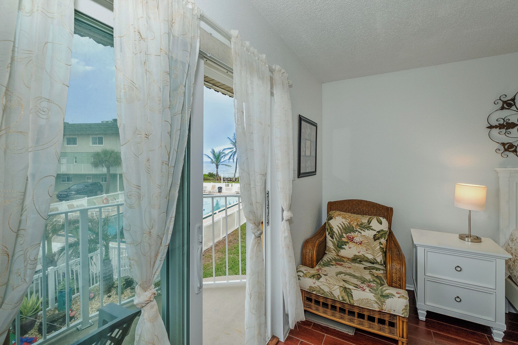 A balcony with a view of a residential area, a wicker chair, a nightstand with a lamp, and a framed picture on the wall.