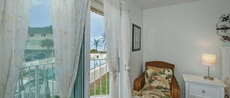 A balcony with a view of a residential area, a wicker chair, a nightstand with a lamp, and a framed picture on the wall.