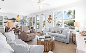 Ein großzügiger Wohnraum mit Sofa, Sessel und Couchtisch, großen Fenstern mit Rollläden sowie einem Deckenventilator.