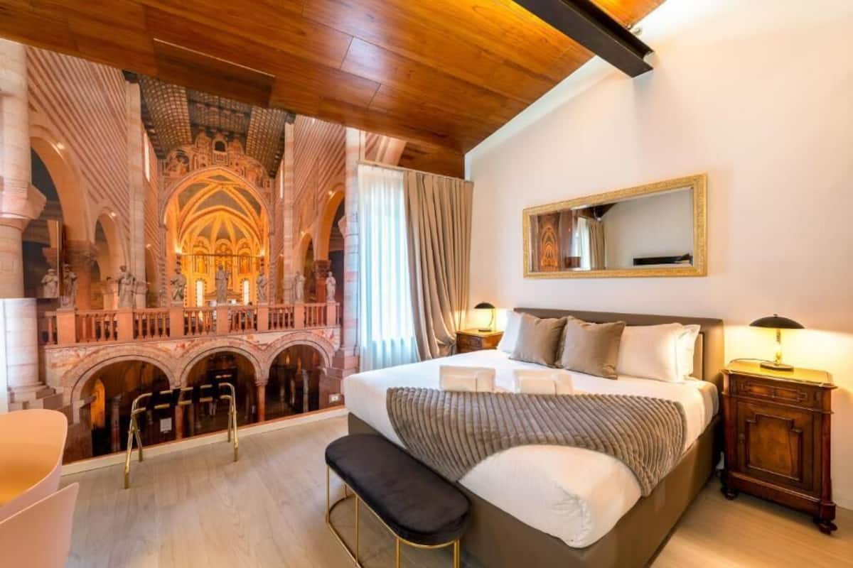 A hotel room with a large bed, a bench, a nightstand, a mirror, and a view of an ornate architectural structure.