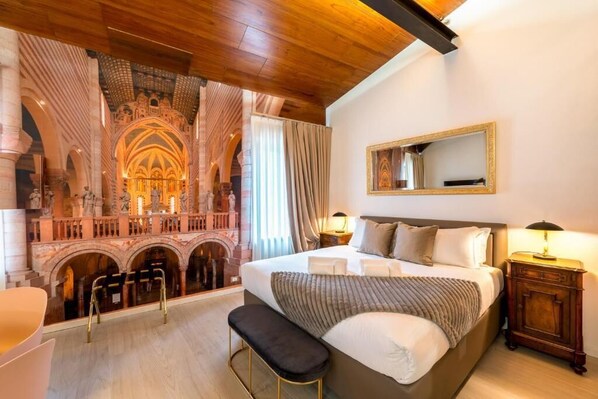 Habitación de hotel con una cama grande, un banco, una mesita de noche, un espejo y vistas a una estructura arquitectónica ornamentada.