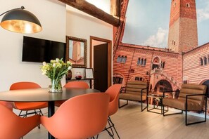 A room with a round table, orange chairs, a vase with flowers, a flat-screen TV, and a mural of a red brick building. - Palazzo Ristori (Verona)