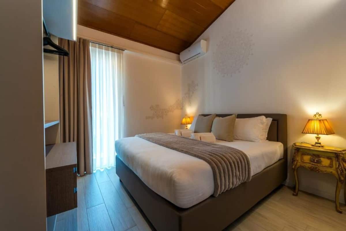 A bedroom with a large bed, bedside lamps, a wooden ceiling, and a window with curtains.